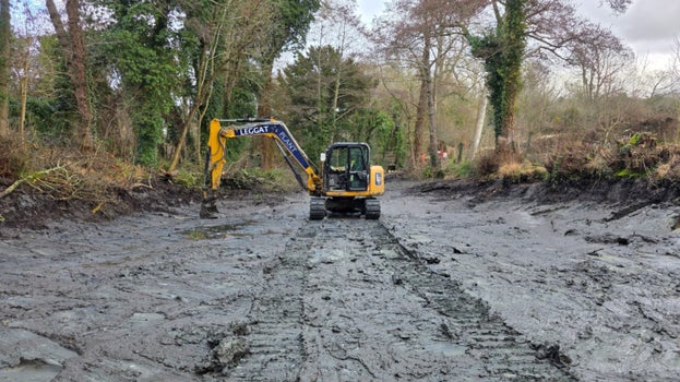 Image of an excavator working on the dark grey mud at the bottom of the drained pond